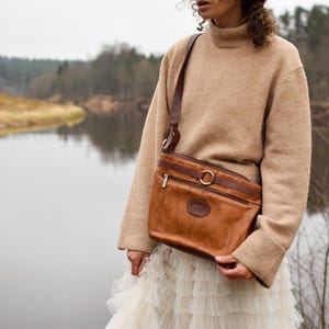May include: A brown leather crossbody bag with a shoulder strap, a zippered top, and a circular metal accent. The bag has a small embossed emblem and is worn over a tan sweater and white skirt.