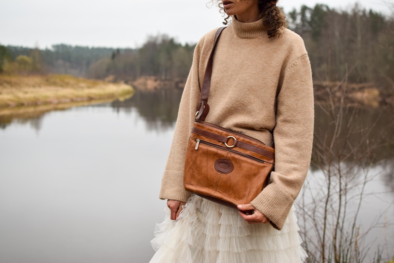 May include: A brown leather crossbody bag with a shoulder strap, a zippered top, and a circular metal accent. The bag has a small embossed emblem and is worn over a tan sweater and white skirt.