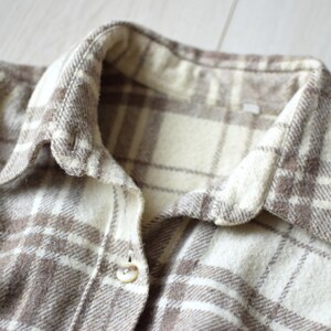 Könnte beinhalten: Nahaufnahme eines beige-braunen karierten Hemdes. Das Hemd hat einen klassischen Kragen und einen Knopfverschluss. Der Stoff hat ein weiches, strukturiertes Aussehen mit einem Muster aus sich kreuzenden Linien.