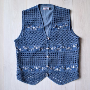 Könnte beinhalten: Eine blaue Jeansweste mit weißen gestickten Blumenmustern und silbernen Knöpfen.