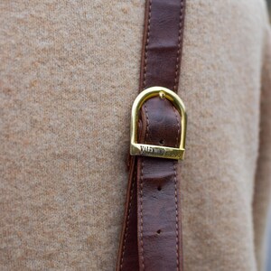 May include: Close-up of a brown leather strap with a gold-colored buckle. The buckle is rectangular with rounded edges and the text "VALENTINO di paolo" is engraved on it. The strap is stitched and appears to be part of a bag or accessory.