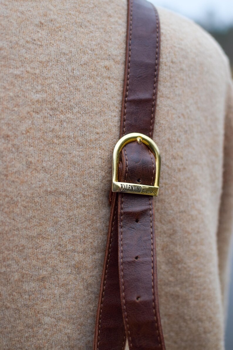 May include: Close-up of a brown leather strap with a gold-colored buckle. The buckle is rectangular with rounded edges and the text "VALENTINO di paolo" is engraved on it. The strap is stitched and appears to be part of a bag or accessory.