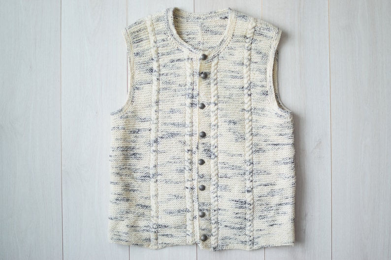 May include: A white knitted vest with a cable knit pattern and button closures. The vest has a subtle gray fleck throughout the fabric.