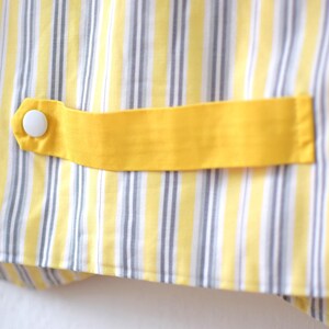 May include: Close-up of a yellow and white striped fabric with a yellow accent. A yellow fabric tab is attached with a white button. The fabric has vertical stripes of yellow, white, and gray.