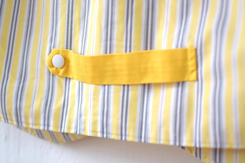 May include: Close-up of a yellow and white striped fabric with a yellow accent. A yellow fabric tab is attached with a white button. The fabric has vertical stripes of yellow, white, and gray.