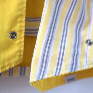 May include: Close-up of a yellow garment with a contrasting striped fabric. The yellow fabric has a smooth texture and metal snap closures. The striped fabric features vertical stripes in yellow, white, and gray.
