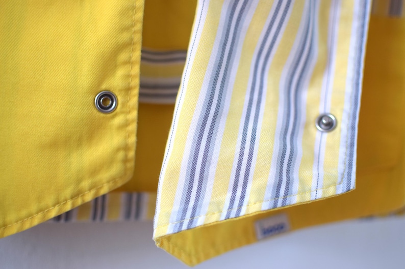 May include: Close-up of a yellow garment with a contrasting striped fabric. The yellow fabric has a smooth texture and metal snap closures. The striped fabric features vertical stripes in yellow, white, and gray.