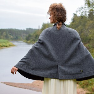 May include: A gray wool cape with a rounded hem and a small pocket on the back. The cape is worn by a person standing in front of a river and trees.