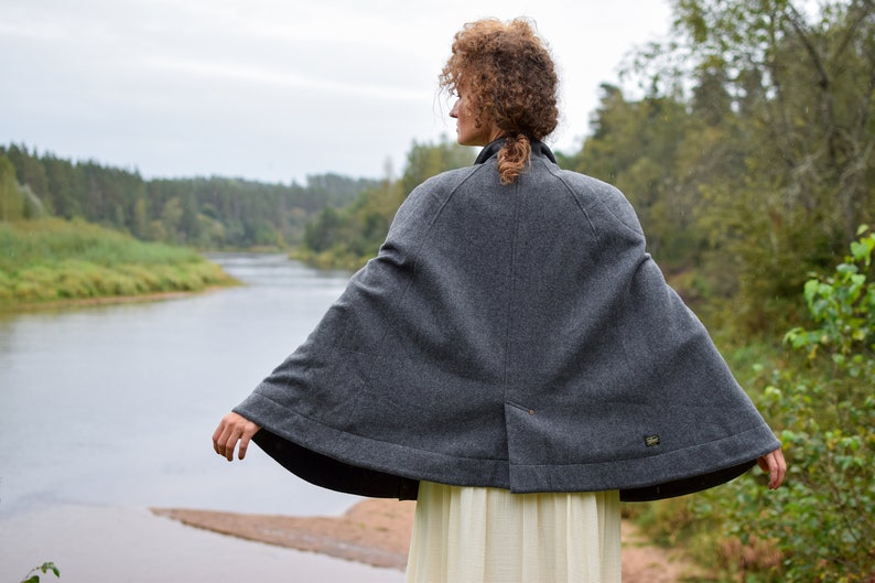 May include: A gray wool cape with a rounded hem and a small pocket on the back. The cape is worn by a person standing in front of a river and trees.