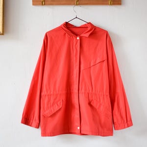 Könnte beinhalten: Eine leuchtend rote Langarmjacke mit Kragen und zwei Vordertaschen. Die Jacke hat einen Knopfverschluss und eine Kordelzug-Taille. Sie hängt an einem schwarzen Kleiderbügel.