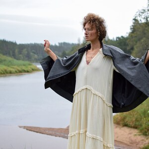 May include: A woman wearing a white dress and a gray cape stands with her arms outstretched in a field near a river. The sky is overcast and the trees in the background are green.