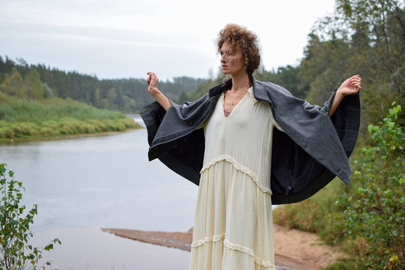 May include: A woman wearing a white dress and a gray cape stands with her arms outstretched in a field near a river. The sky is overcast and the trees in the background are green.