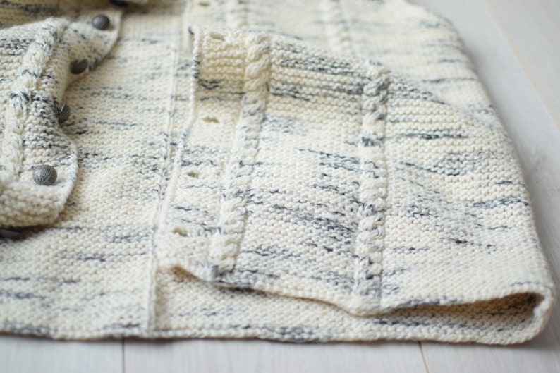May include: A white and gray knitted cardigan with a button closure and a cable knit detail on the front.