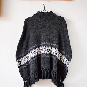 Könnte beinhalten: Ein dunkelgraues Poncho mit hohem Kragen und einem weißen und grauen geometrischen Muster auf der Brust. Das Poncho hat Fransen am unteren Rand und hängt an einer Holzgarderobe.