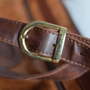 May include: Close-up of a brown leather bag strap with a gold-colored buckle. The buckle is rectangular and has the words "VALENTINO di paolo" engraved on it. The leather has visible stitching.