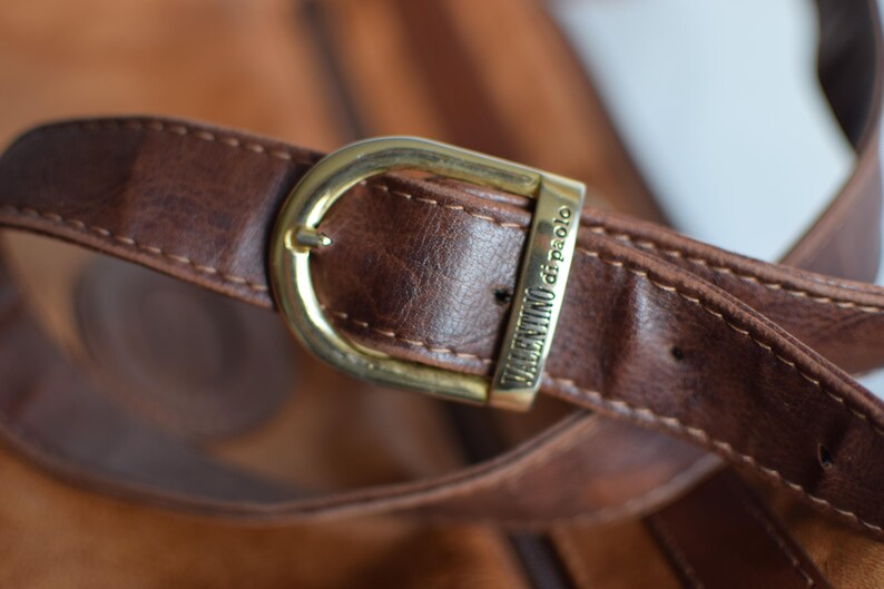 May include: Close-up of a brown leather bag strap with a gold-colored buckle. The buckle is rectangular and has the words "VALENTINO di paolo" engraved on it. The leather has visible stitching.