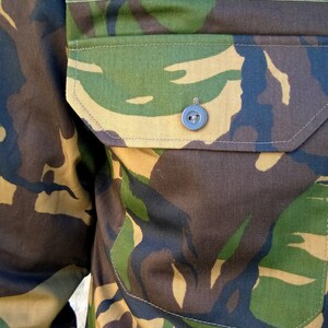 May include: A close-up of a camouflage fabric with a green velcro patch and a button. The fabric has a brown, green, and tan pattern.