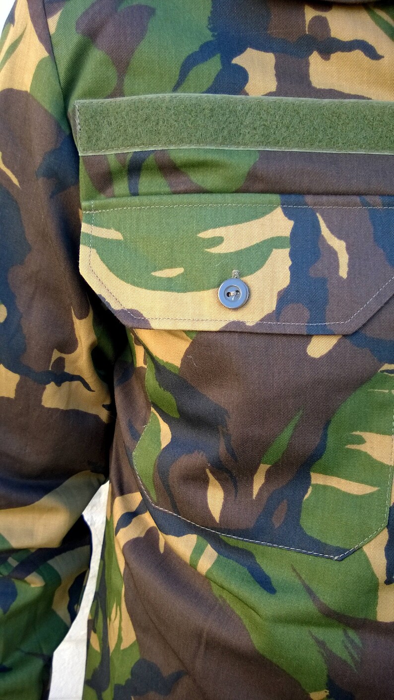May include: A close-up of a camouflage fabric with a green velcro patch and a button. The fabric has a brown, green, and tan pattern.