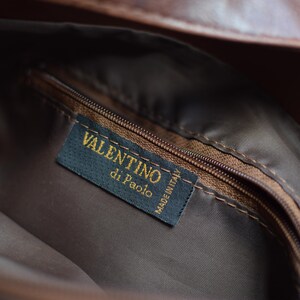 May include: Close-up of the interior of a brown leather bag. A dark blue label with gold text reads "VALENTINO di Paolo MADE IN ITALY". The bag has a brown zipper and a brown fabric lining.