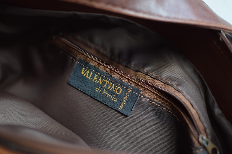 May include: Close-up of the interior of a brown leather bag. A dark blue label with gold text reads "VALENTINO di Paolo MADE IN ITALY". The bag has a brown zipper and a brown fabric lining.