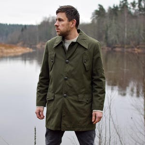 May include: A man wearing a green, long-sleeved, button-up jacket with a collar. He is standing in front of a body of water with trees in the background.