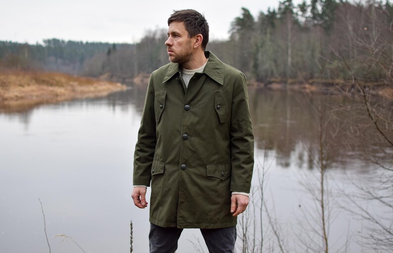 May include: A man wearing a green, long-sleeved, button-up jacket with a collar. He is standing in front of a body of water with trees in the background.