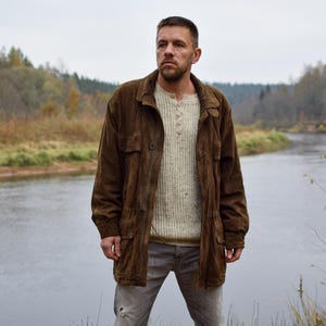 Könnte beinhalten: Eine Person trägt eine braune Wildlederjacke über einem cremefarbenen Pullover und grauen Hosen in der Nähe eines Flusses. Die Jacke hat einen Knopfverschluss und mehrere Taschen. Der Hintergrund zeigt einen Fluss und Bäume.