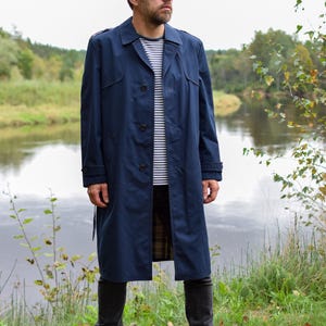 Könnte beinhalten: Ein marineblauer Trenchcoat mit klassischem Kragen, Knopfleiste und Schulterklappen. Der Mantel wird über einem weiß-marineblau gestreiften T-Shirt, einer dunkelgrauen Hose und schwarzen Schnürstiefeln aus Leder getragen.