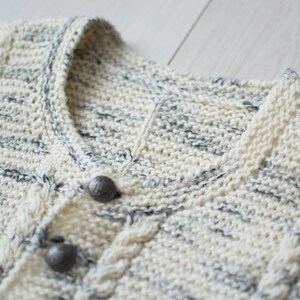 May include: Close-up of a cream-colored knitted cardigan with a cable knit pattern and two silver buttons. The cardigan has a subtle gray and white speckled pattern.