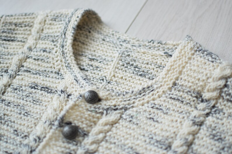 May include: Close-up of a cream-colored knitted cardigan with a cable knit pattern and two silver buttons. The cardigan has a subtle gray and white speckled pattern.