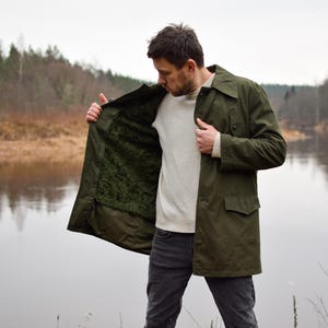 May include: A man wearing a green jacket with a brown fleece lining. He is standing by a river and looking down at the jacket.
