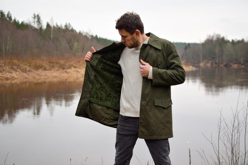 May include: A man wearing a green jacket with a brown fleece lining. He is standing by a river and looking down at the jacket.