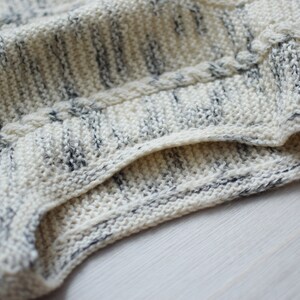 May include: Close-up of a white and gray knitted fabric with a cable knit pattern. The fabric is folded over, showing the inside of the knit.