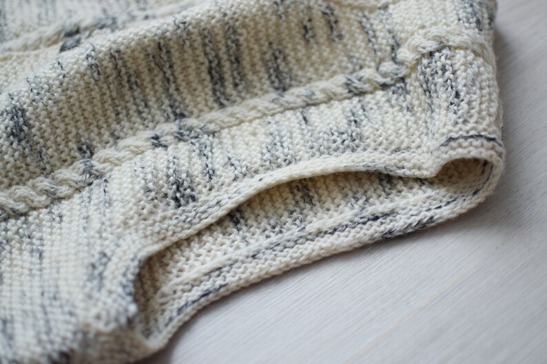 May include: Close-up of a white and gray knitted fabric with a cable knit pattern. The fabric is folded over, showing the inside of the knit.