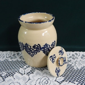 May include: A cream-colored ceramic jar with a blue and white floral design. The jar has a lid with a knob on top. The jar is sitting on a white lace tablecloth.