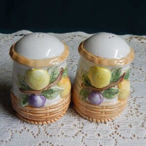 May include: A set of two ceramic salt and pepper shakers with a fruit and basket design. The shakers are white with a brown basket design and feature yellow lemons, purple plums, and green leaves.