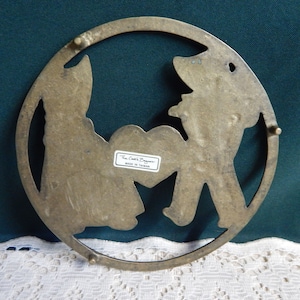May include: Brass trivet with a cutout design of two figures holding a heart. The trivet is round and has a label that reads "The Cook's Bazaar Made in Taiwan".