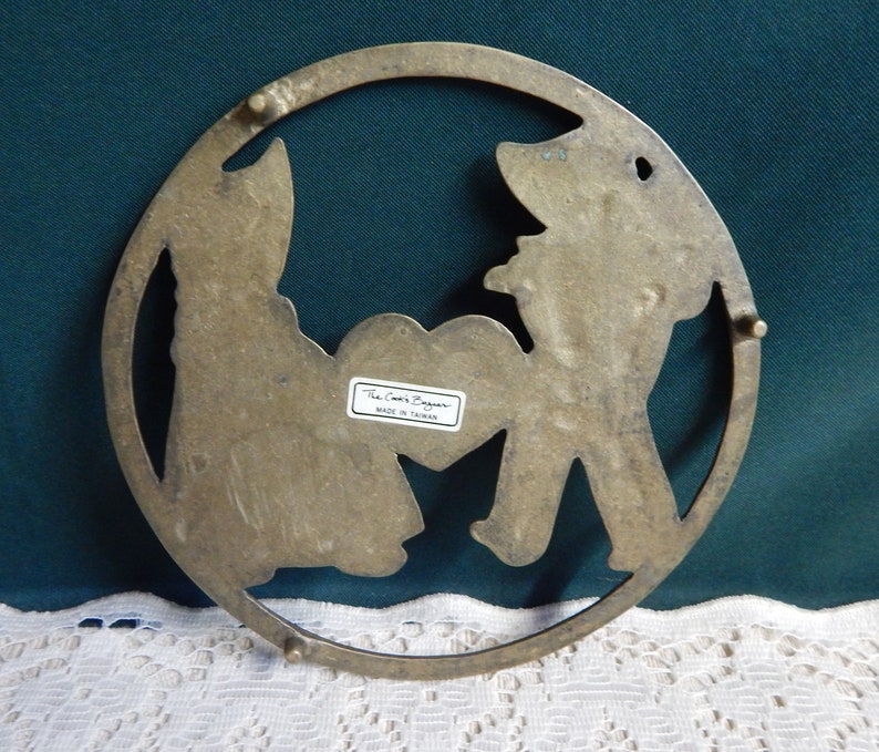 May include: Brass trivet with a cutout design of two figures holding a heart. The trivet is round and has a label that reads "The Cook's Bazaar Made in Taiwan".