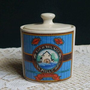 May include: A cylindrical ceramic canister with a lid, featuring a blue vertical striped background and a central circular graphic depicting a mill house with a water wheel. The text reads Oak Wood Mills Grainery Established 1859, Premium Feed Seed.