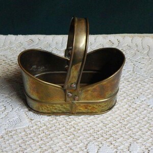 May include: An oval-shaped brass basket with a polished metallic finish and a single arched handle. The basket features a smooth, rounded body with visible rivets securing the handle to the sides, showcasing a warm, golden-brown patina.