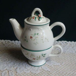 May include: A white ceramic teapot and teacup set. The teapot has a lid with a handle and is decorated with green and red floral accents. The teacup is stacked beneath the teapot, with a matching floral design and a green trim.