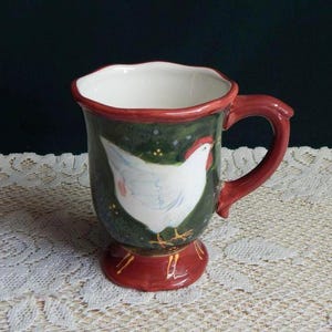 May include: A red and green ceramic mug with a white chicken design. The mug has a footed base and a handle.