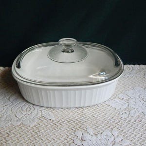 May include: An oval white ceramic casserole dish with vertical ribbed sides and a clear glass lid. The lid features a central circular knob handle surrounded by a radial sunburst pattern etched into the glass surface, providing a clear view.