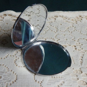 May include: Open, oval-shaped silver compact mirror. One side is a standard mirror, the other has a mirror and a dark green powder area. The compact is open, revealing the mirrors and powder.