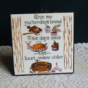 May include: A square white ceramic tile with a dark metal frame featuring brown and purple illustrations of food and drink. The text reads: Give me yesterdays bread, This days meat, and last years cider. It includes drawings of bread, meat, honey, butter, and a cider barrel.