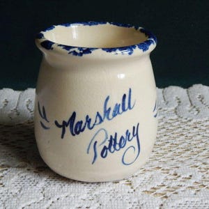 May include: Off-white ceramic jar with a rounded body and a slightly flared rim. The rim is decorated with blue brushstrokes. The words "Marshall Pottery" are written in blue script on the front of the jar.
