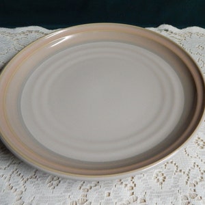 May include: A large, round, ceramic dinner plate with a light beige color and a multi-colored rim. The rim has a light brown, beige, and pink color.