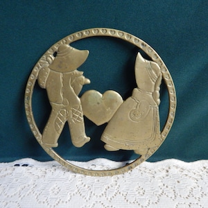May include: A brass trivet with a cutout design of two figures holding a heart. The figures are dressed in traditional clothing and are surrounded by a circle of hearts.