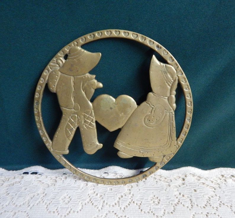 May include: A brass trivet with a cutout design of two figures holding a heart. The figures are dressed in traditional clothing and are surrounded by a circle of hearts.