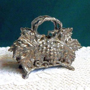 May include: Silver napkin holder with a grapevine design. The holder is made of metal and has a detailed design of leaves and grapes.